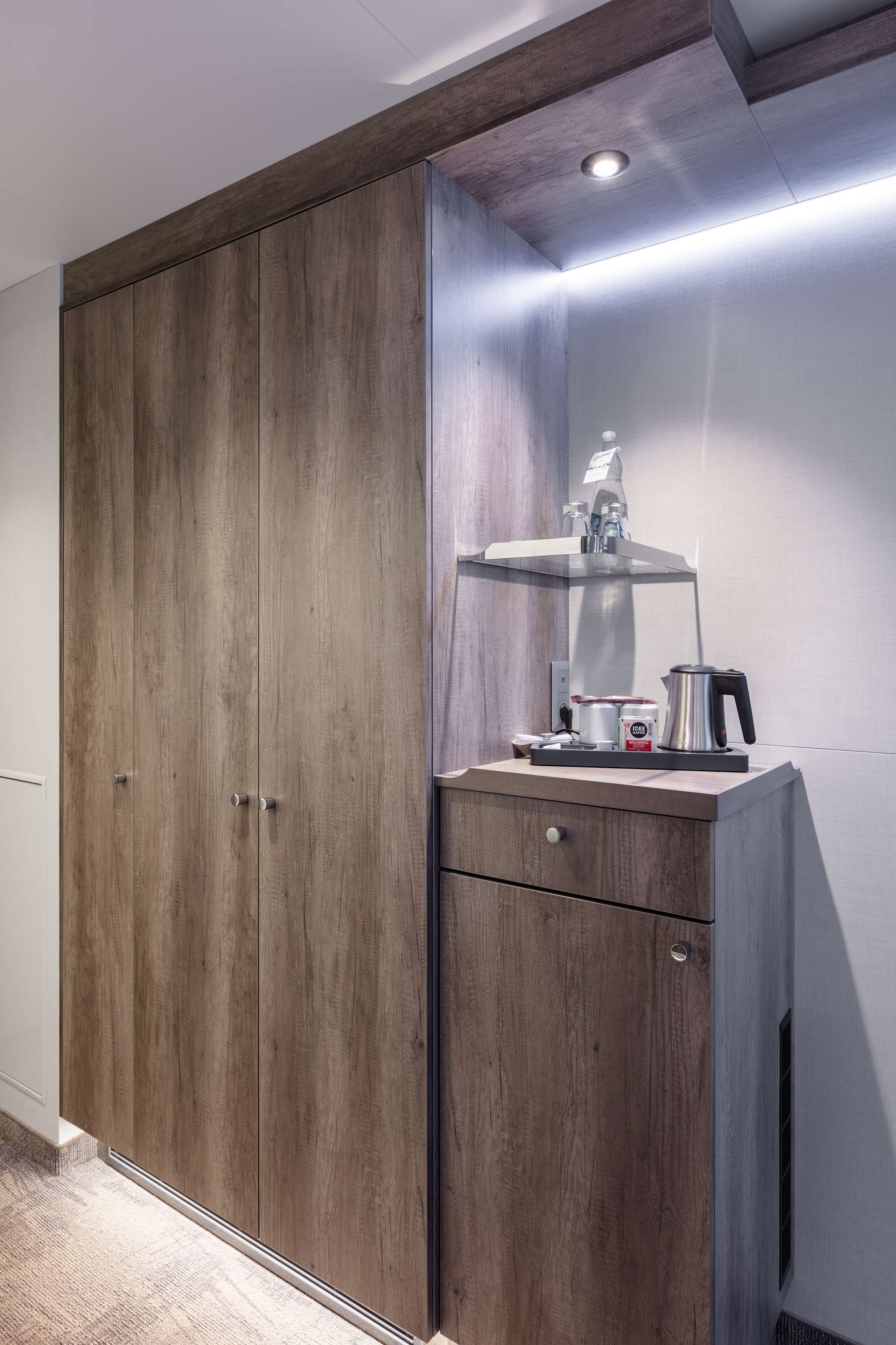Ein Schrank mit Holzoptik, eine kleine Ablagefläche mit Wasserkocher und Kaffeemaschine sowie ein an der Wand befestigtes Regal.
