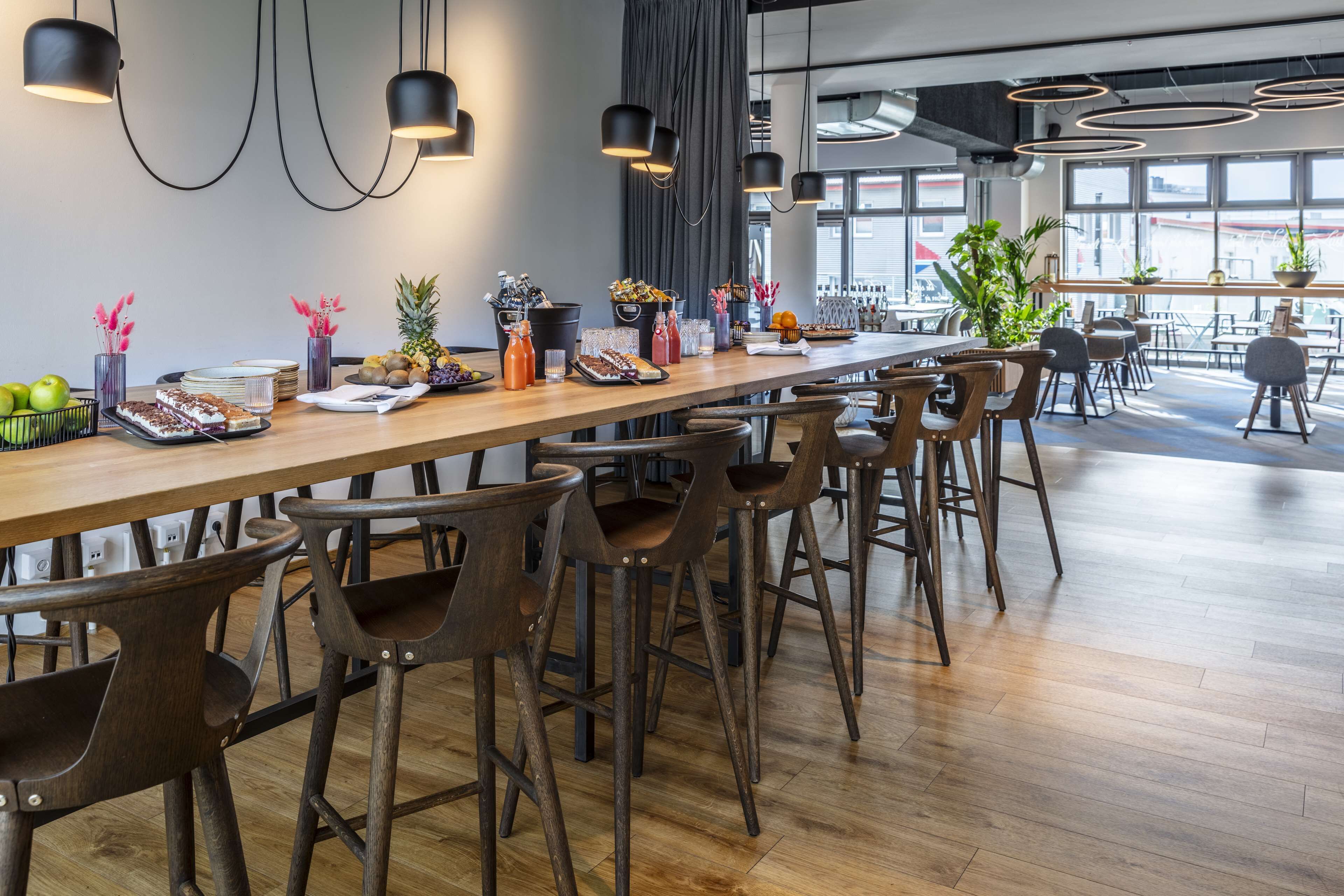 Un espace de restauration moderne avec une longue table en bois, des tabourets de bar et des luminaires suspendus.