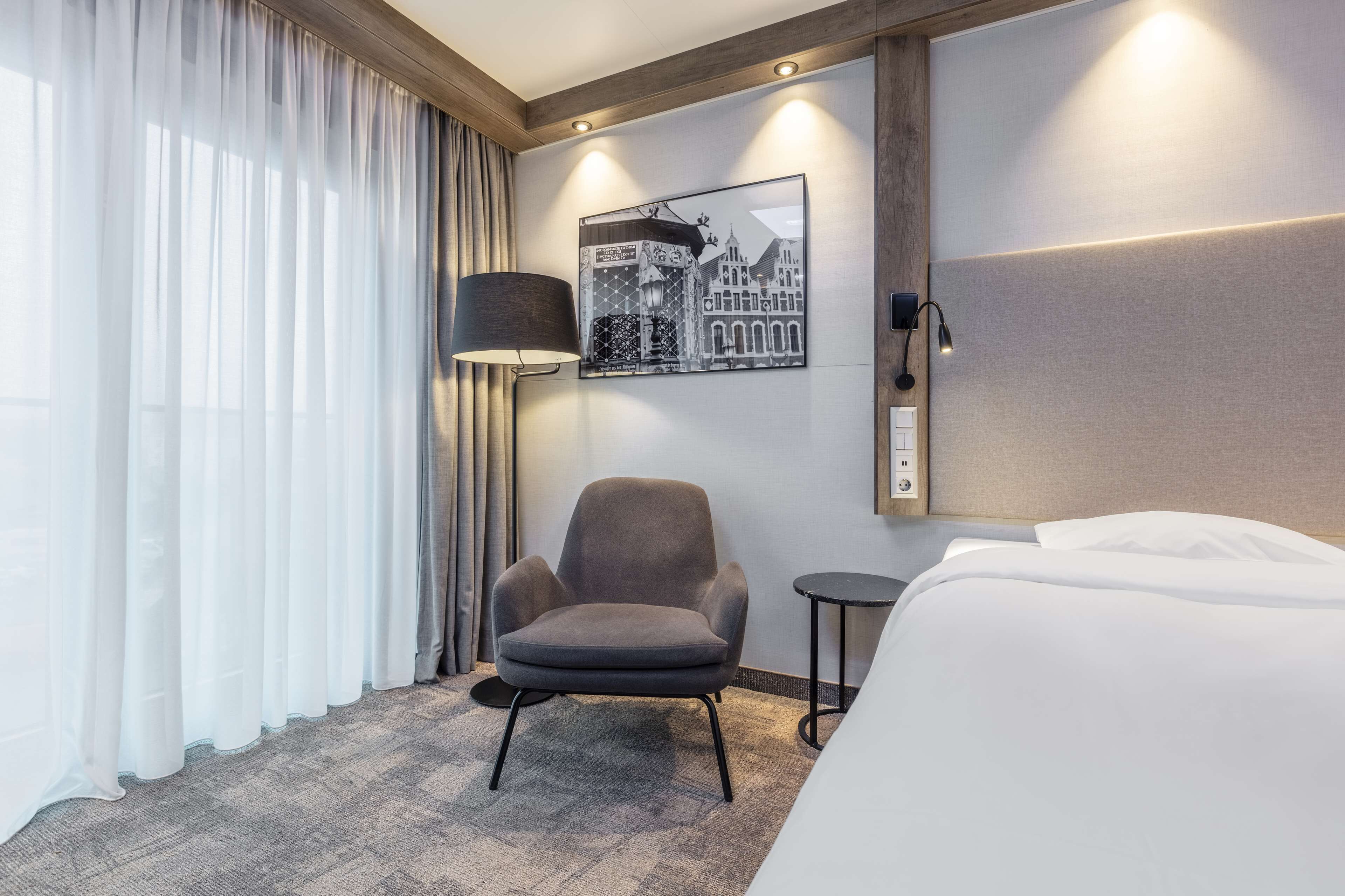 Une chambre d’hôtel avec un lit, une chaise, une lampe, un tableau au mur et une fenêtre avec des rideaux.