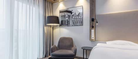 Una habitación de hotel con cama, una silla, lámpara, un cuadro en la pared y una ventana con cortinas.
