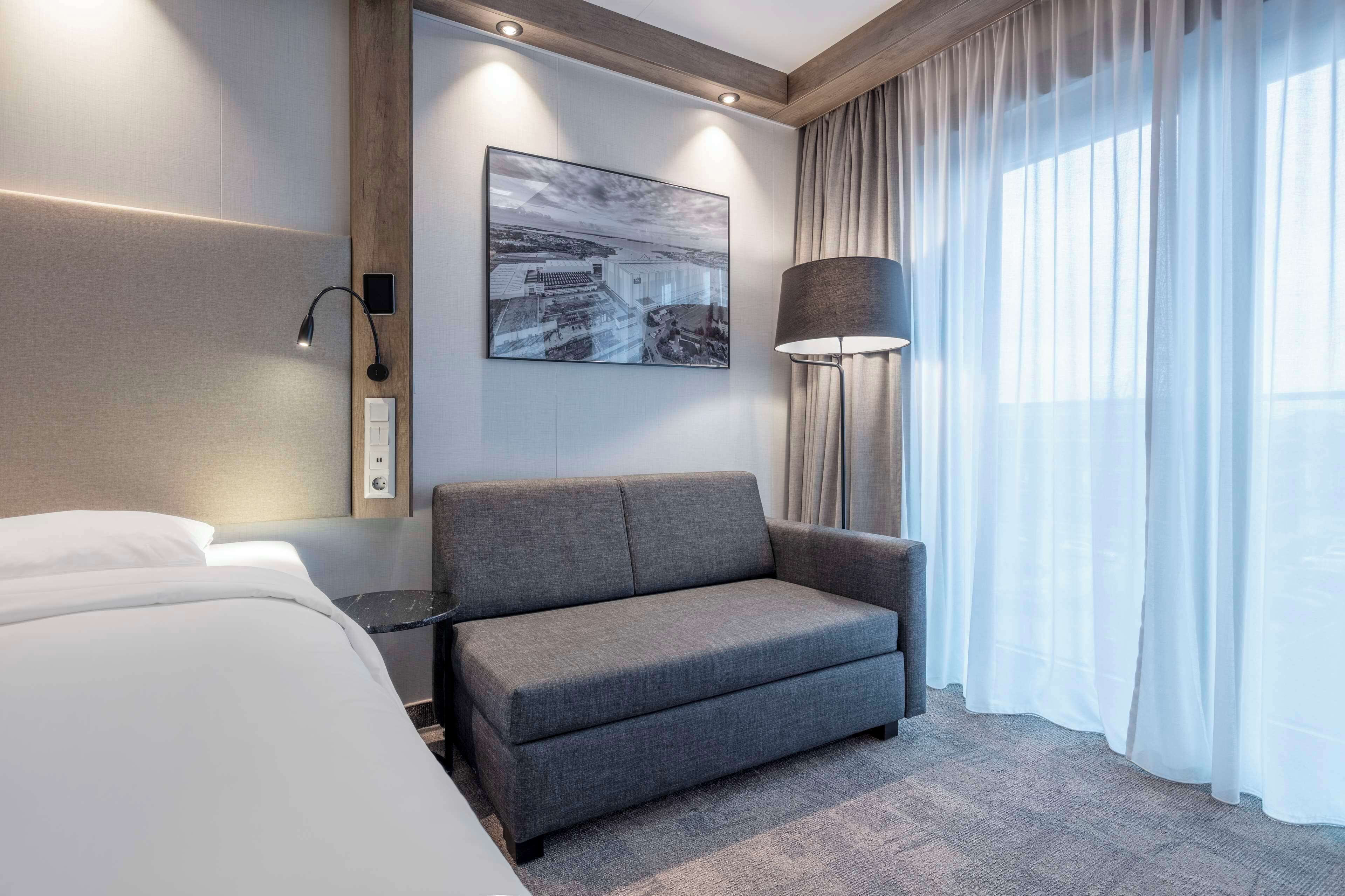 A modern hotel room with a grey sofa, a large window with sheer curtains, a wall-mounted lamp, and a framed cityscape artwork.