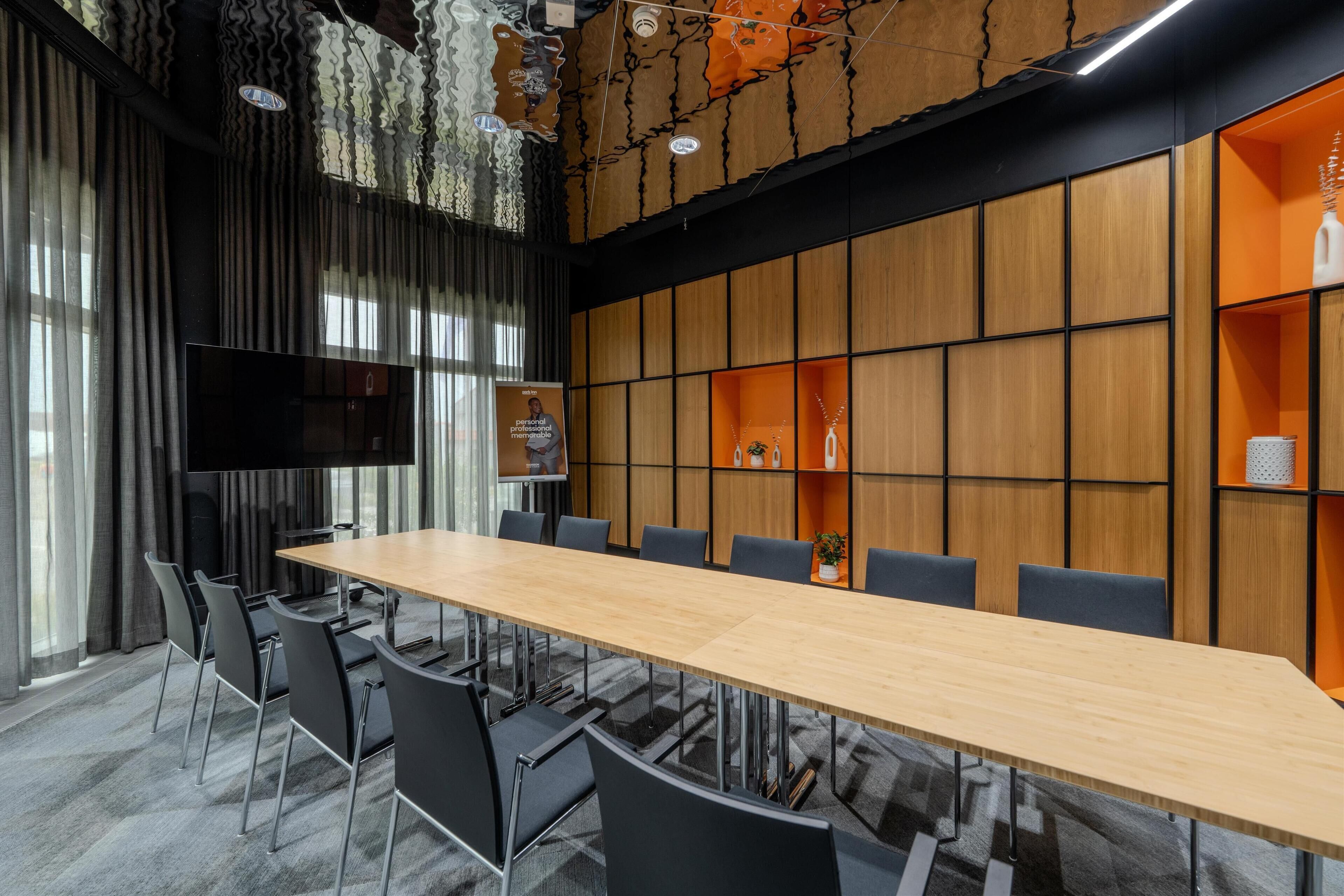 Ein Konferenzraum mit einem langen Tisch, Stühlen, einem Fernseher und holzverkleideten Wänden mit orangefarbenen Regalen.