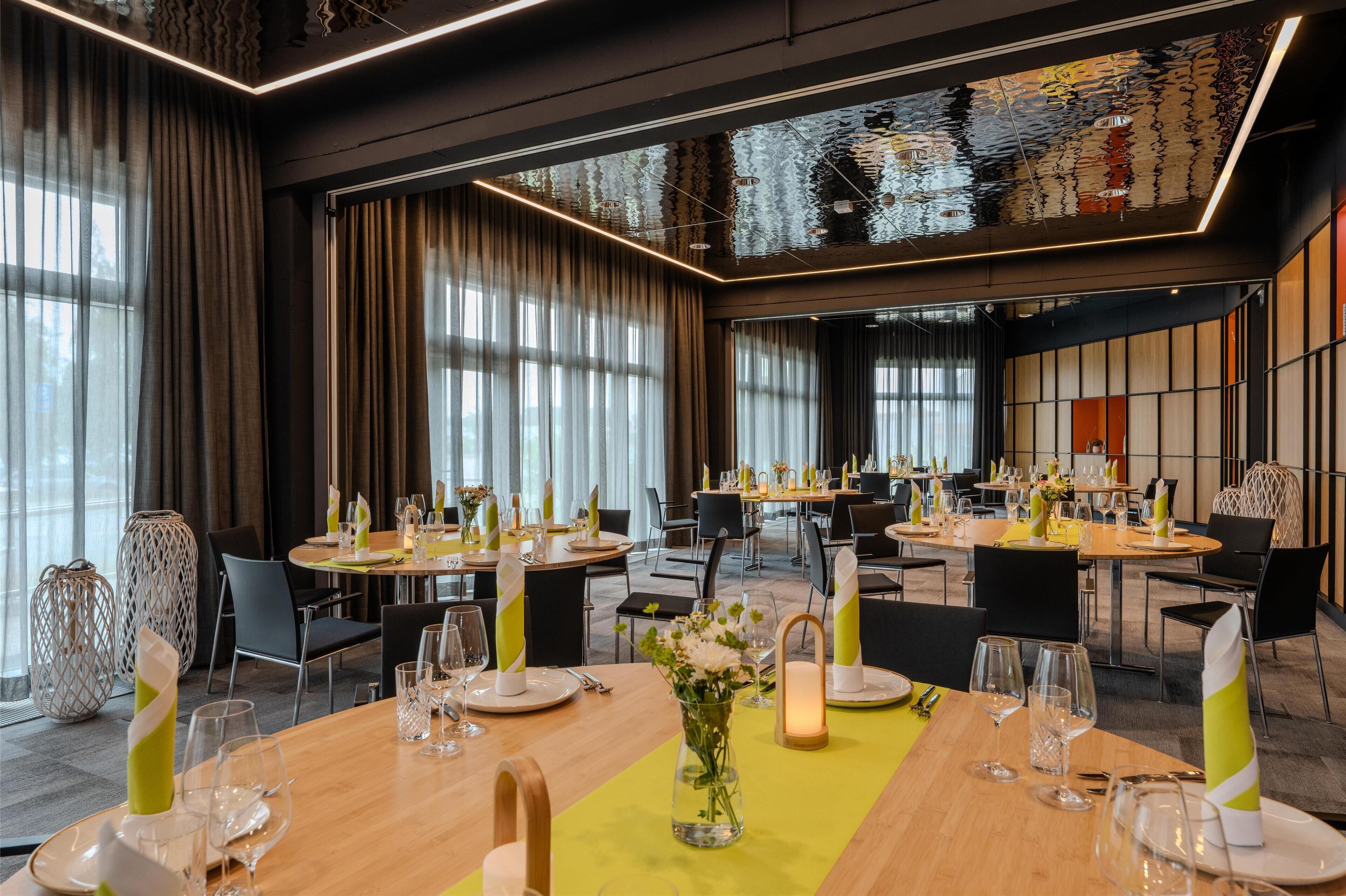 A modern dining area with round tables set for a meal, large windows with curtains, and a unique ceiling design reflecting light.
