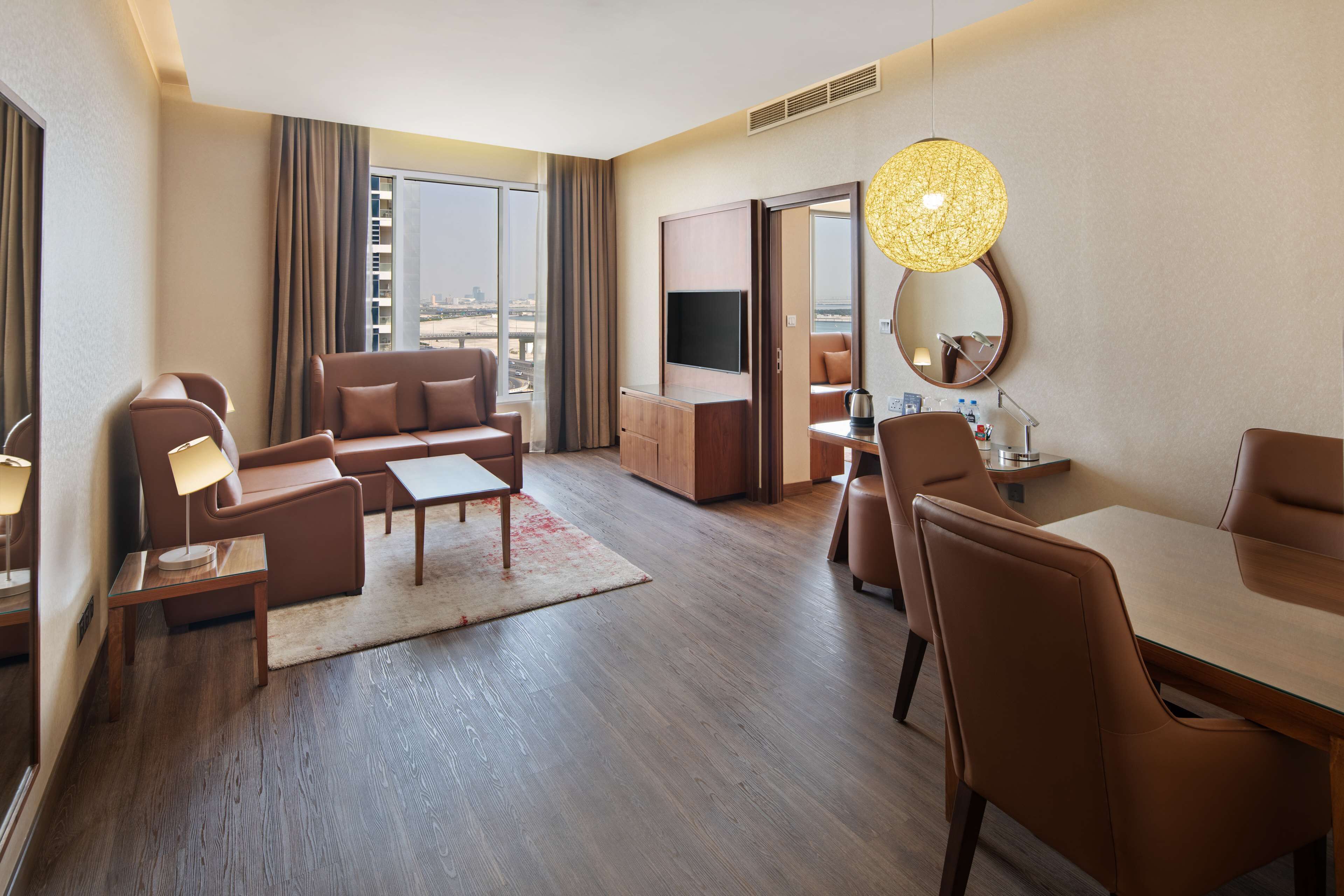 Et moderne hotelværelse med sofa, sofabord, spisebord og stole. Der er et fjernsyn, et spejl og en lampe.