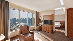 A modern hotel room with a large window offering a city view, a comfortable seating area, a flat-screen TV, and a neatly made bed.