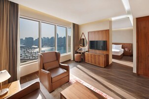 A modern hotel room with a large window offering a city view, a comfortable seating area, a flat-screen TV, and a neatly made bed.