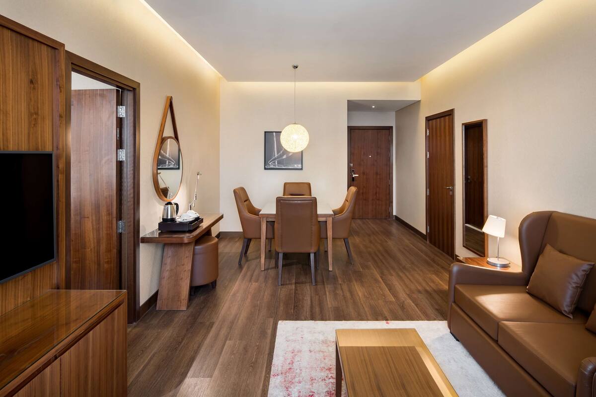 A modern living room with a wooden floor, a brown sofa, a dining table with chairs, a TV, and a lamp.