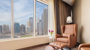 A modern hotel room with a large window offering a city view, a brown leather armchair, a small round table with a vase of flowers, and a white floor lamp.