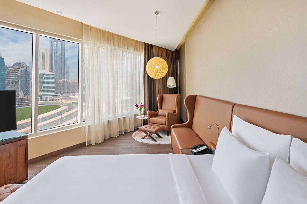 A hotel room with a large bed, a brown armchair, a small side table, a lamp, and a view of a cityscape through large windows.
