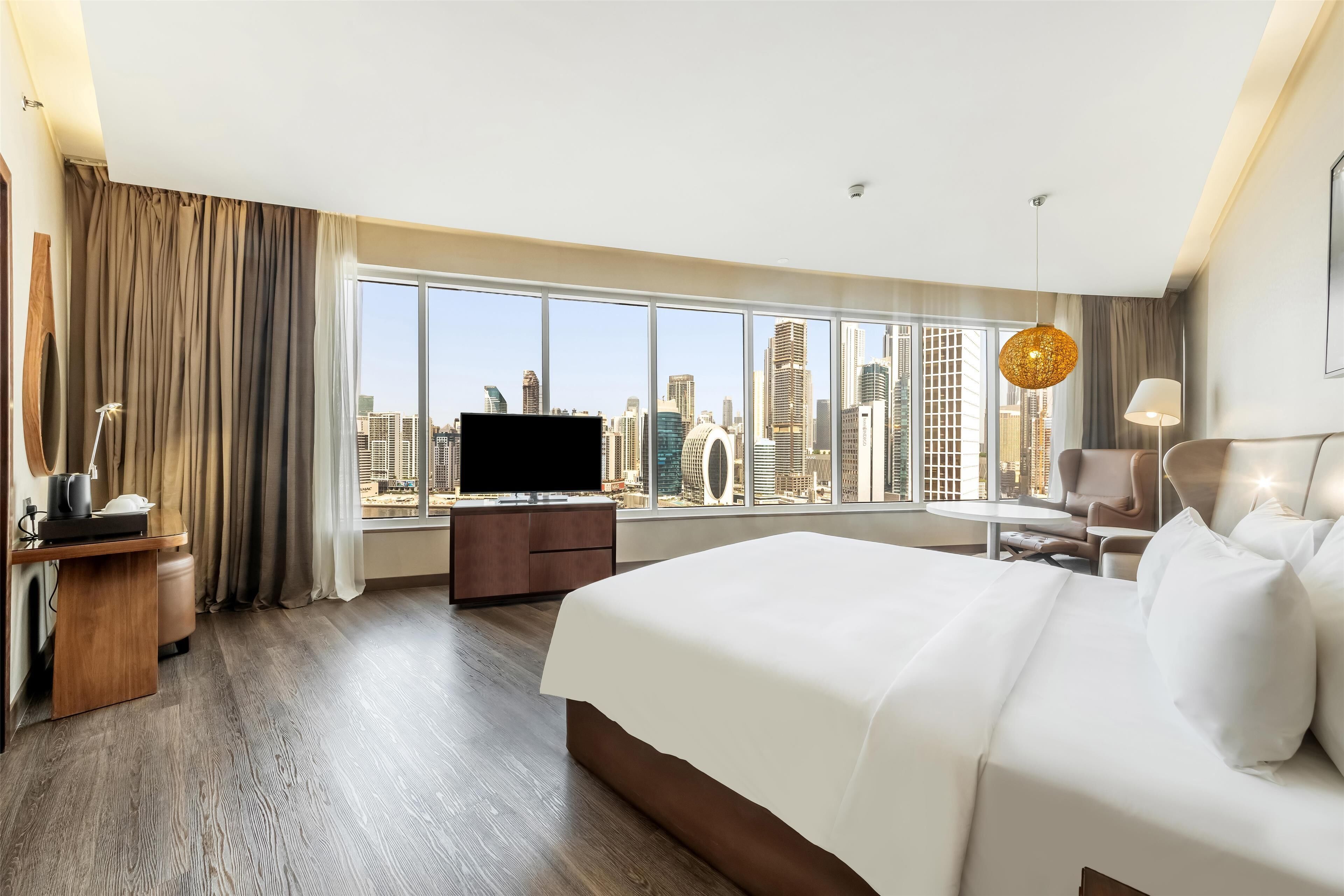 Ein Hotelzimmer mit einem großen Bett, einem Schreibtisch, einem Flachbildfernseher, einem Sessel und Blick auf eine Stadtlandschaft durch große Fenster.