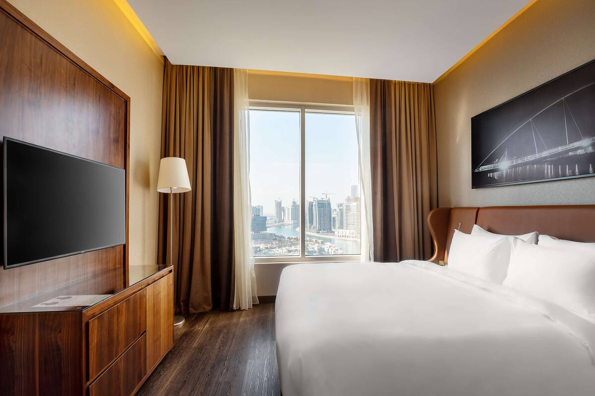 A hotel room with a large bed, a flat-screen TV, a wooden cabinet, a lamp, and a window with a city view.