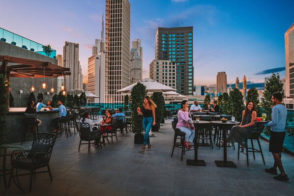 An outdoor dining area with tables and chairs, surrounded by tall buildings and a clear sky.