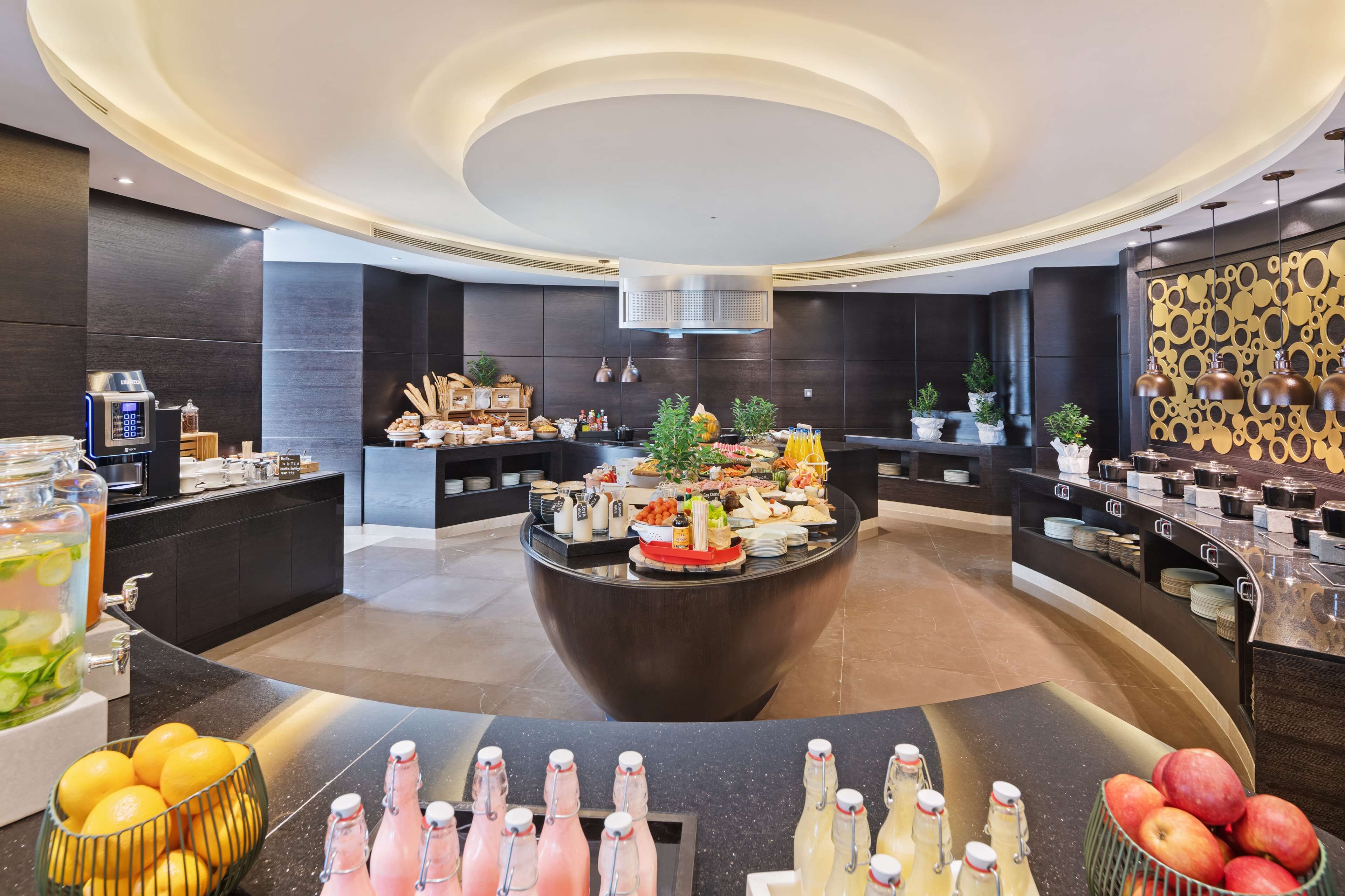 A modern hotel lobby with a central island featuring a buffet, surrounded by dark-toned walls and a circular ceiling design.
