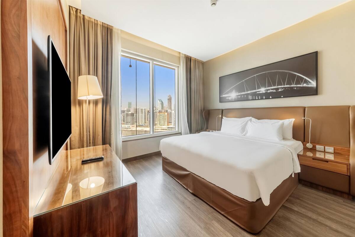 A hotel room with a large bed, a flat-screen TV, a desk with a lamp, and a city view through the window.