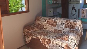 A room with a bed covered in a leopard print blanket, a window with a view of greenery, and a kitchenette with a refrigerator and shelves. - Bangalôs Pico do Avião em frente à IIha do Campeche (Florianópolis)
