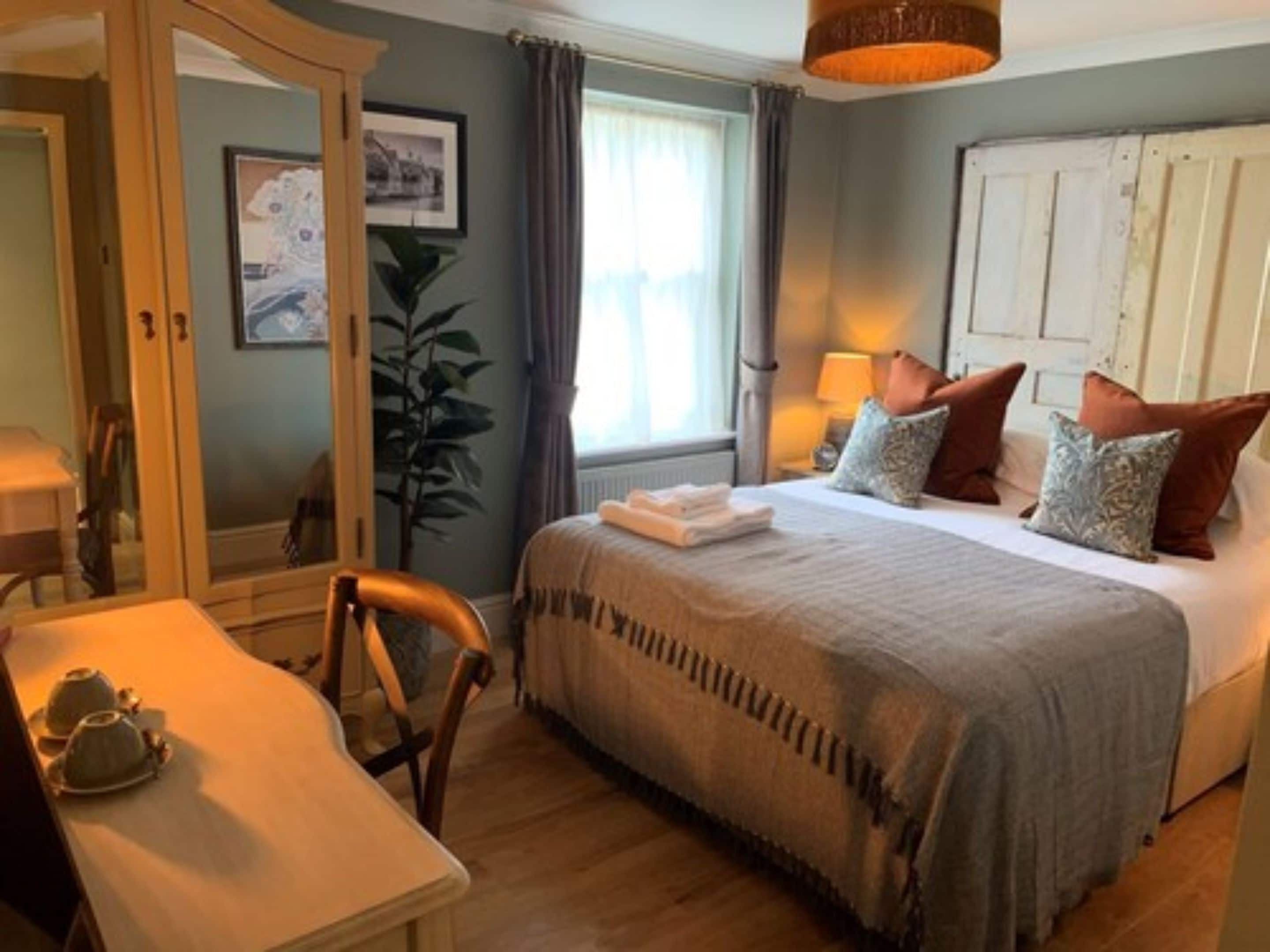 A hotel room with a bed, a desk, a chair, a plant, and a window with curtains.