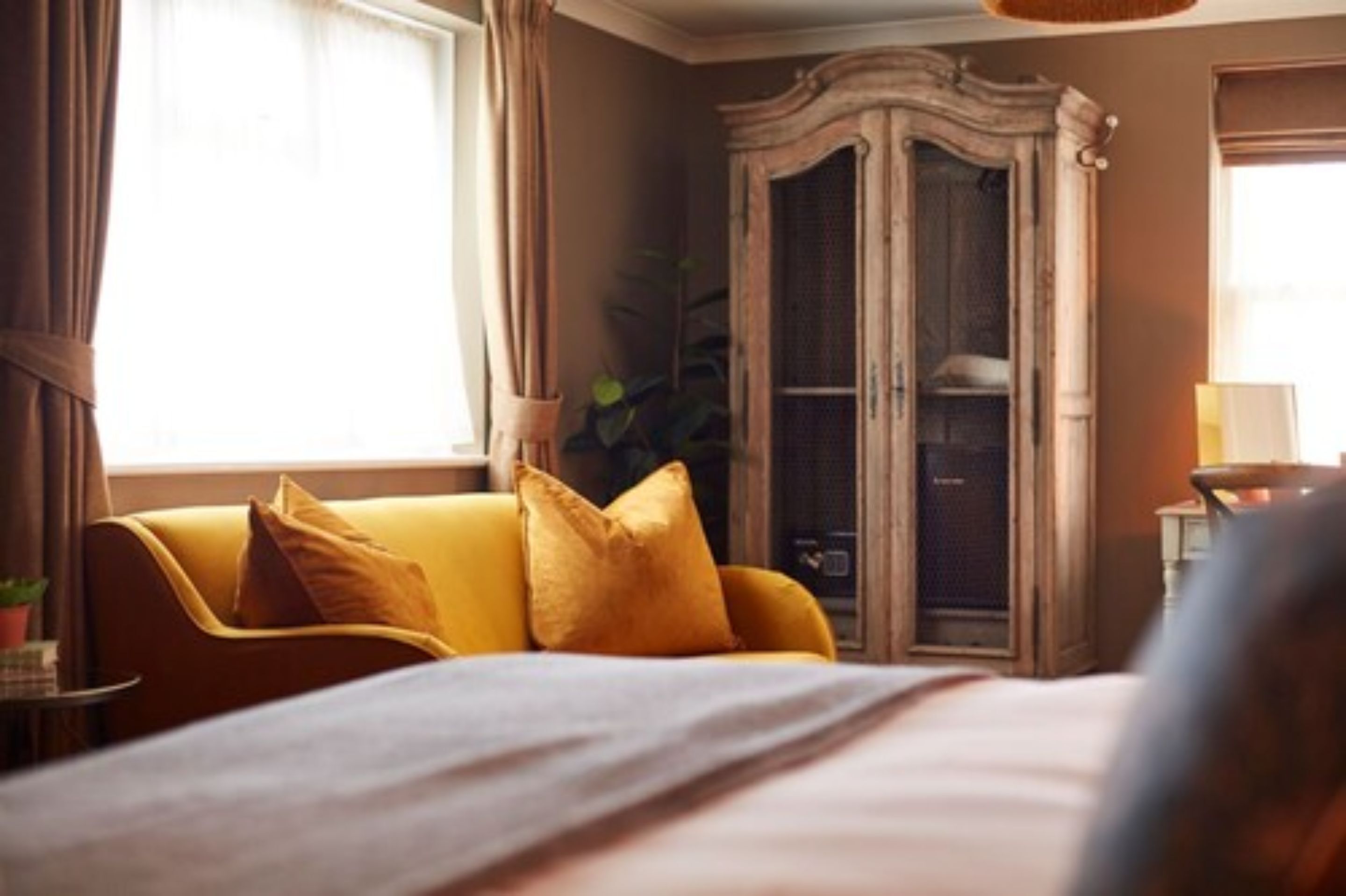 A hotel room with a bed, a yellow sofa, a wooden wardrobe, and a window with curtains.