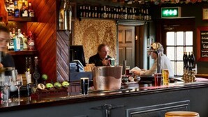 A bar with wooden shelves stocked with bottles, a cash register, and a bar stool.
