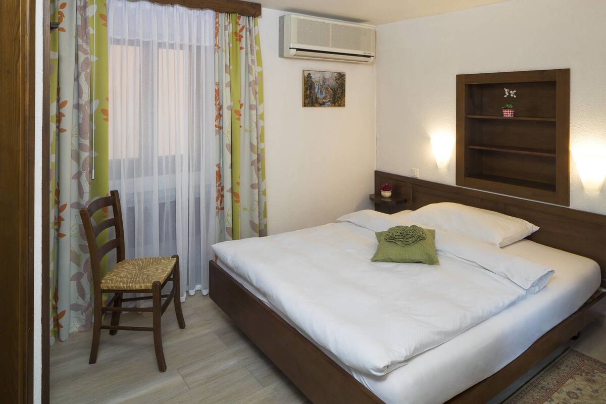 A hotel room with a bed, a chair, an air conditioner, a painting, and a shelf with a plant.
