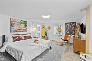 A bedroom with a bed, a chair, a surfboard, and a sign that says 'PRAY FOR SURF'.