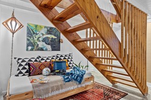 A loft-style bedroom with a wooden staircase, a bed with patterned pillows, a wall-mounted painting, and a hanging light fixture.