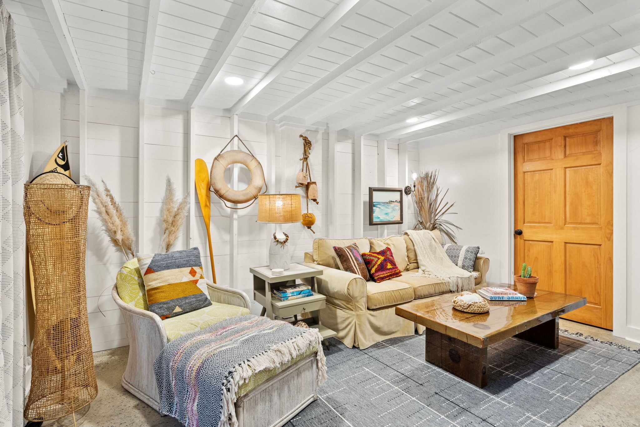 A cozy living room with a white wooden ceiling, a beige sofa, a patterned armchair, a wooden coffee table, and a wicker basket.