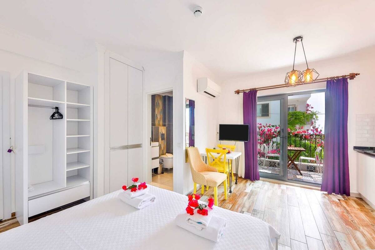 A hotel room with a bed, a wardrobe, a bathroom, a dining area, and a balcony with flowers.