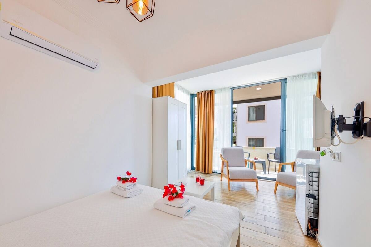 A hotel room with a bed, two chairs, a small table, a TV, and a balcony with curtains.
