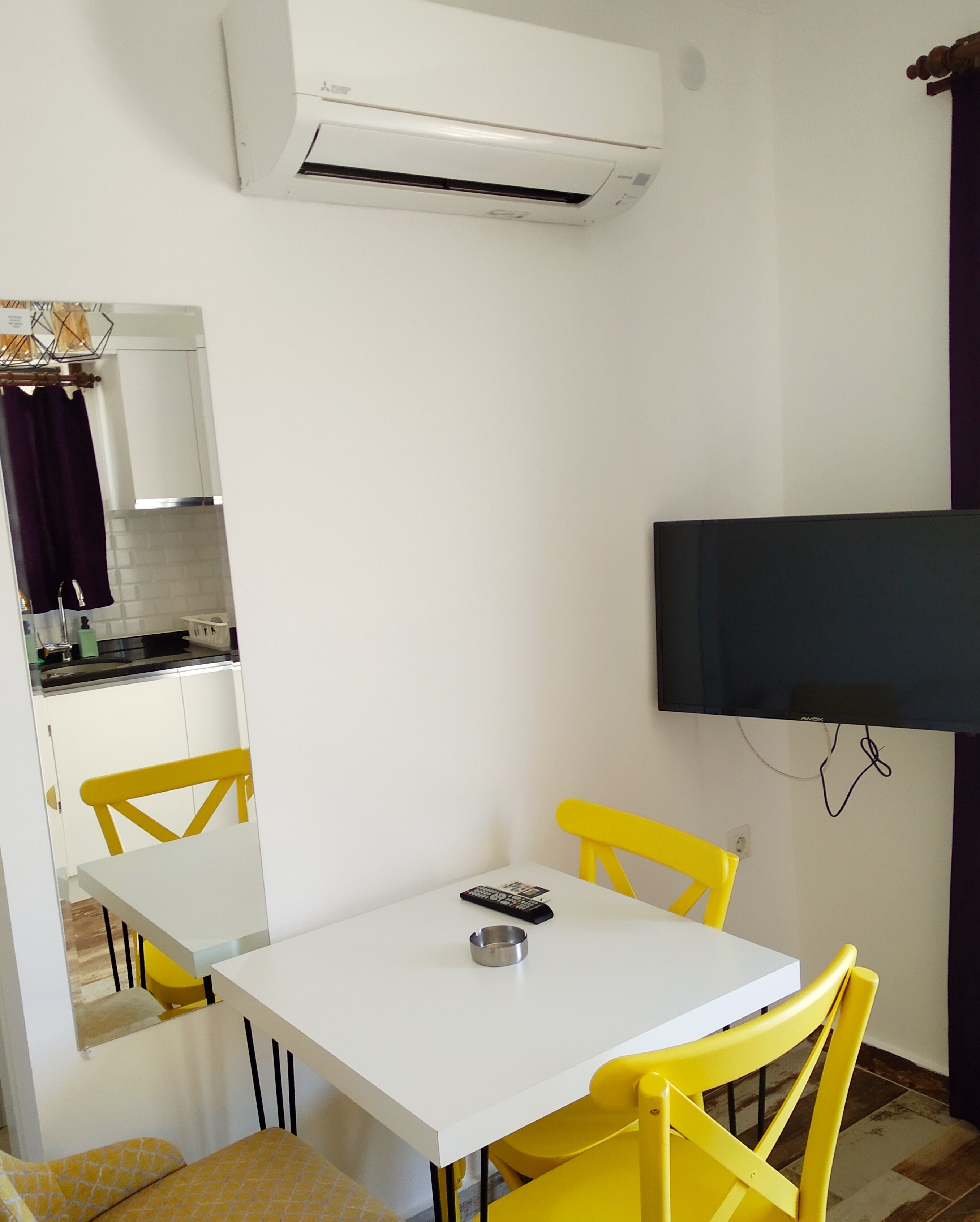 A small, modern room with a white table and yellow chairs, a flat-screen TV mounted on the wall, and a kitchen area visible through an open door.