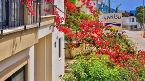 A building with a balcony adorned with red flowers, a sign reading "SANISU APARTS", and a clear view of the street and other buildings in the background. - Kas Sanisu Pansiyon (Kas)