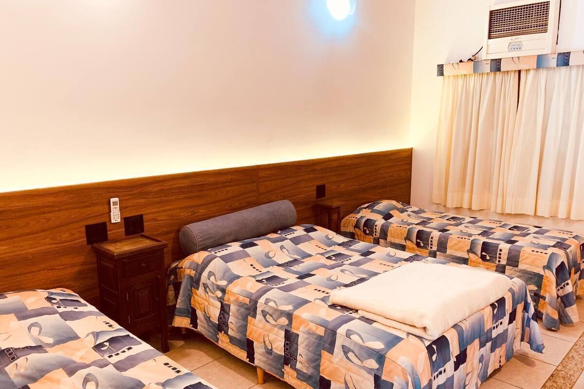 A hotel room with two beds, a wooden headboard, a bedside table, a wall-mounted air conditioner, and a curtain.