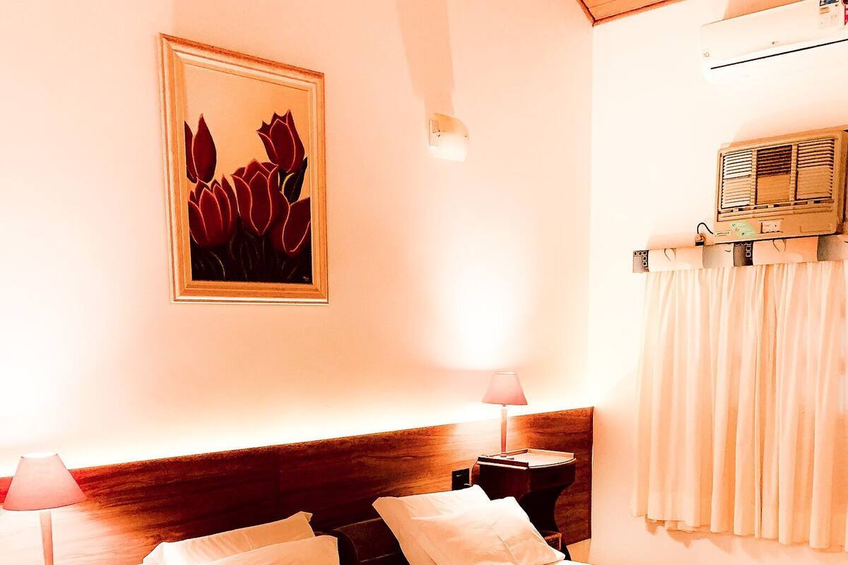 A bedroom with a large bed, bedside tables, a lamp, a wall-mounted air conditioner, and a painting.