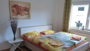 A neatly made bed with a colorful bedspread, a bedside table with a vase, and a window with a view of the outside. - Wohnung im Zentrum des Ruhrgebiets (Castrop-Rauxel)