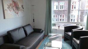 A modern living room with a dark leather sofa, a glass coffee table, and a view of buildings through large windows. - Wohnung im Zentrum des Ruhrgebiets (Castrop-Rauxel)