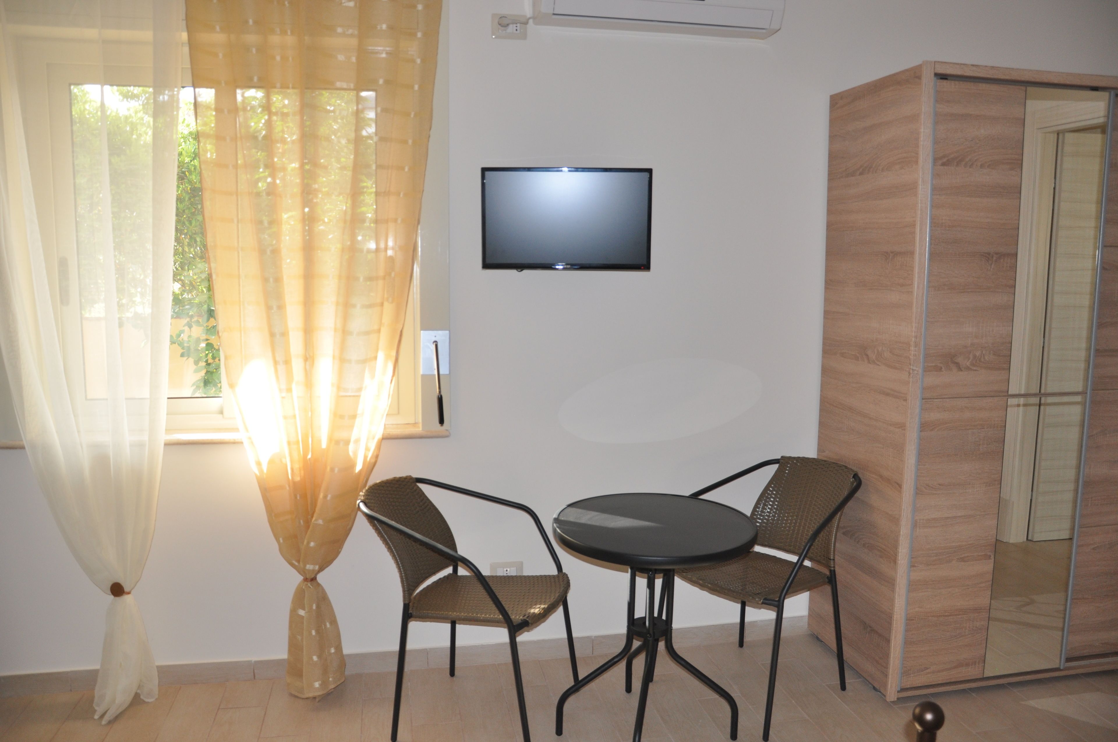 A room with a TV mounted on the wall, a round table with two chairs, and a wardrobe with a mirror.