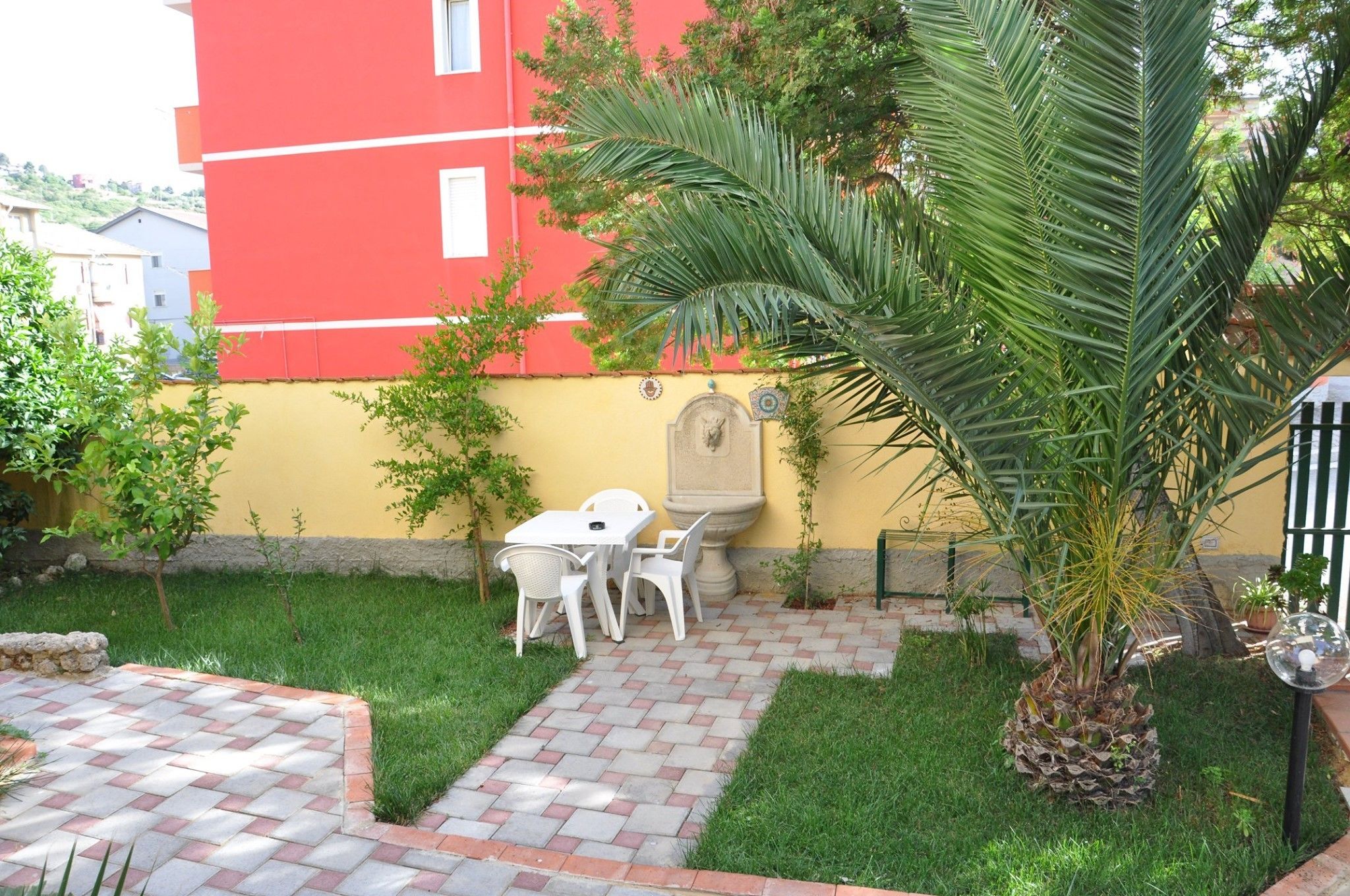 Un patio con solado, una mesa y sillas blancas, una palmera y un edificio rojo al fondo.