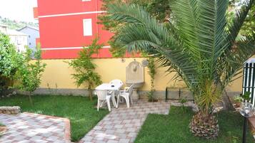 Un patio con solado, una mesa y sillas blancas, una palmera y un edificio rojo al fondo.