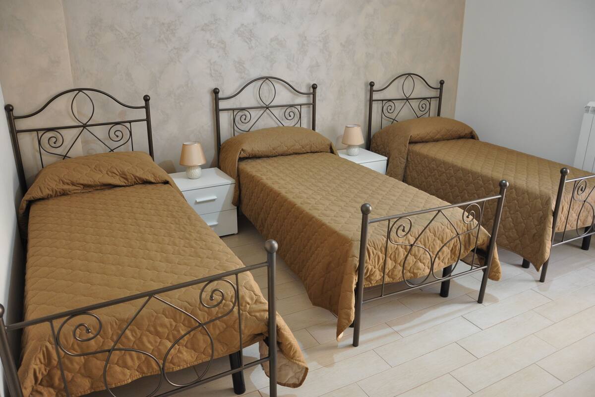 A room with three single beds, each with a metal frame and a brown quilted bedspread. There are bedside tables with lamps and a white wall in the background.
