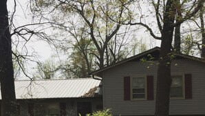 Une maison à deux étages, dotée d’un toit en pente, entourée d’un jardin bien entretenu où poussent diverses plantes et fleurs.