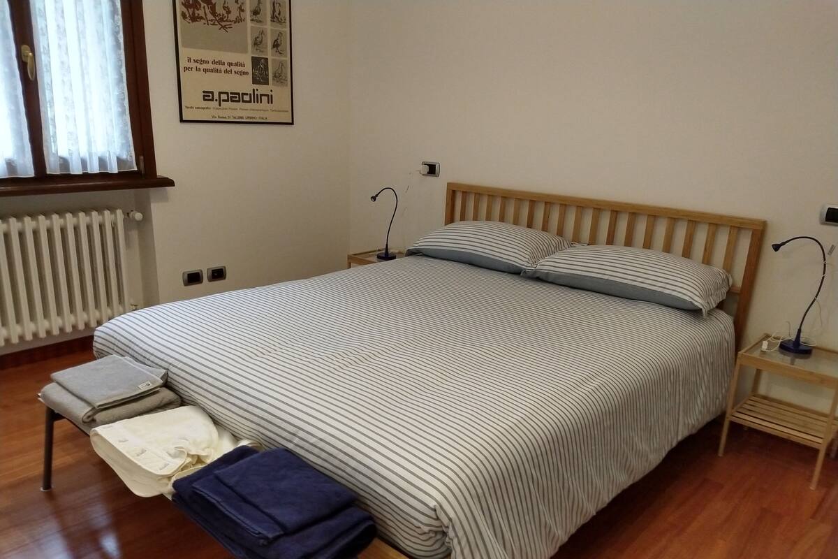 A bedroom with a large bed, a bedside table, a nightstand, a radiator, and a window with curtains.