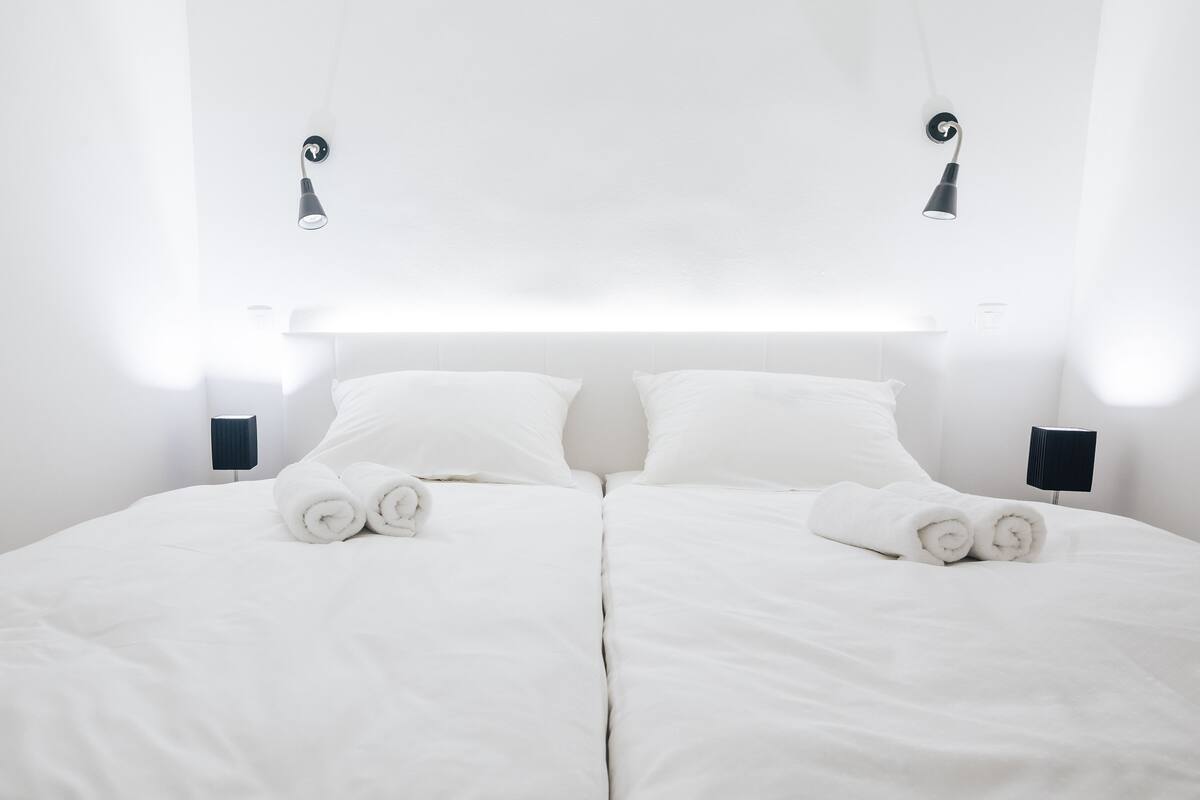 Een groot bed met witte lakens en opgerolde handdoeken, twee nachtkastjes met lampen, en een wit plafond met hangende lampen.