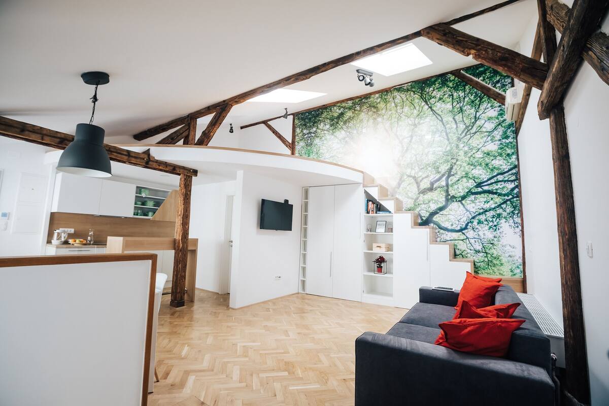 Een modern interieur met een gebogen witte muur, houten balken, een zwarte hanglamp en uitzicht op bomen door een groot raam.