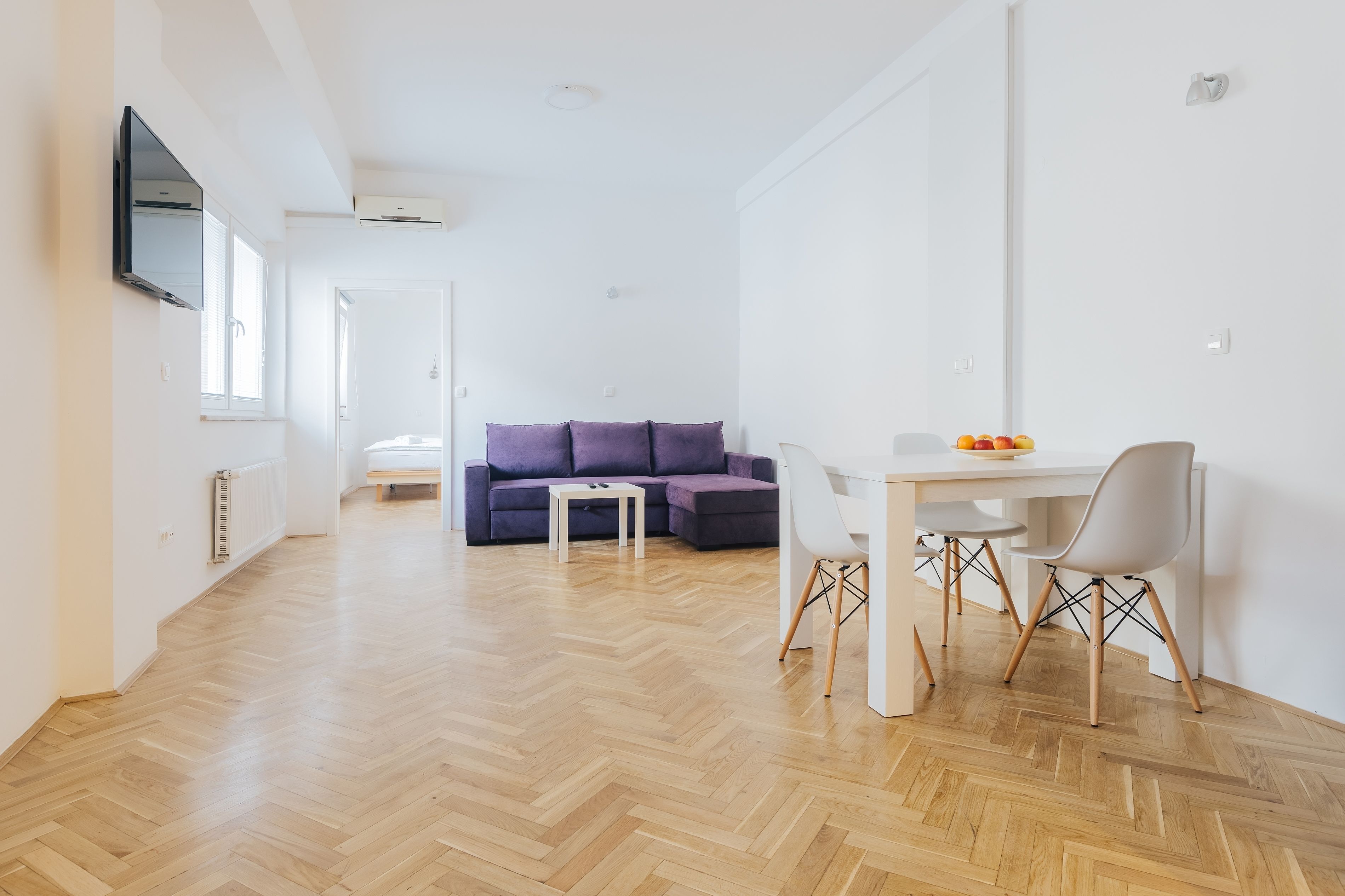 Ein modernes Wohnzimmer mit einer Couch, einem Esstisch und Stühlen. Ein Fernseher ist an der Wand befestigt, und ein Fenster lässt natürliches Licht herein.