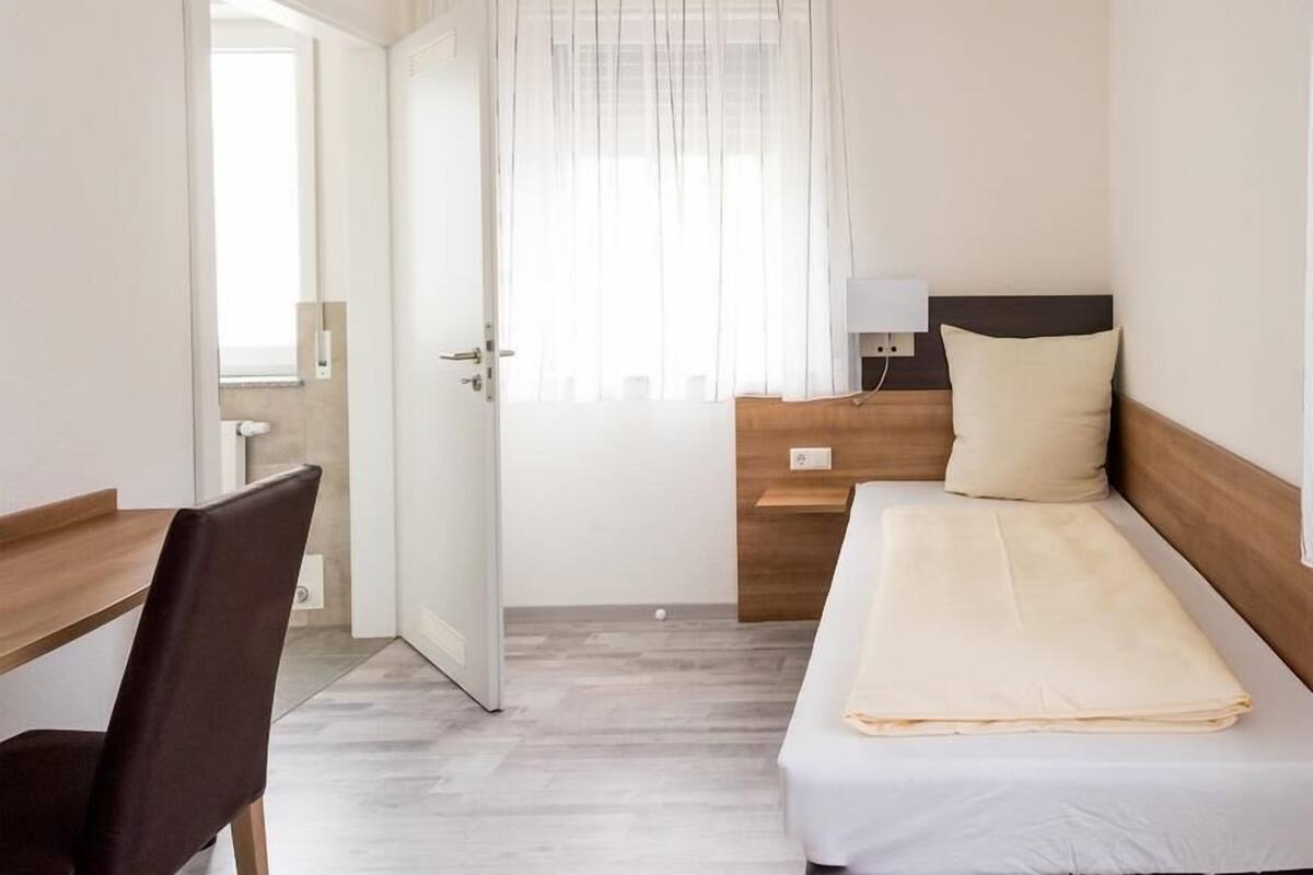 A bedroom with a bed, a desk, and a chair. There is a window with curtains, a bedside table with a lamp, and a door leading to another room.