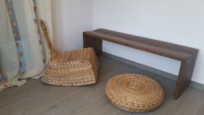 Een rieten stoel en voetenbankje naast een houten bank in een kamer met een betegelde vloer en een witte muur.