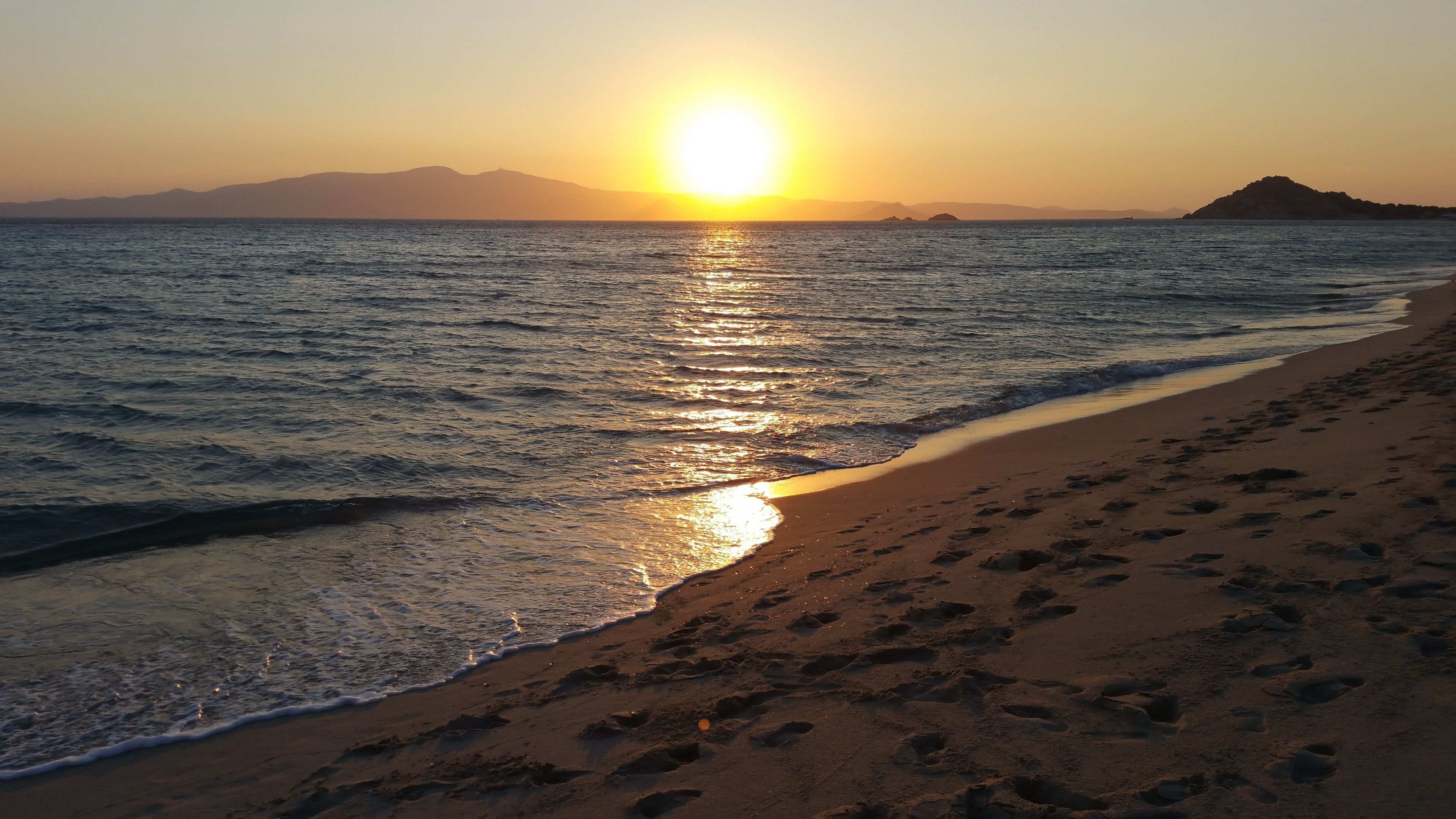 A beach with footprints leading to the water, a sunset in the background, and distant mountains.