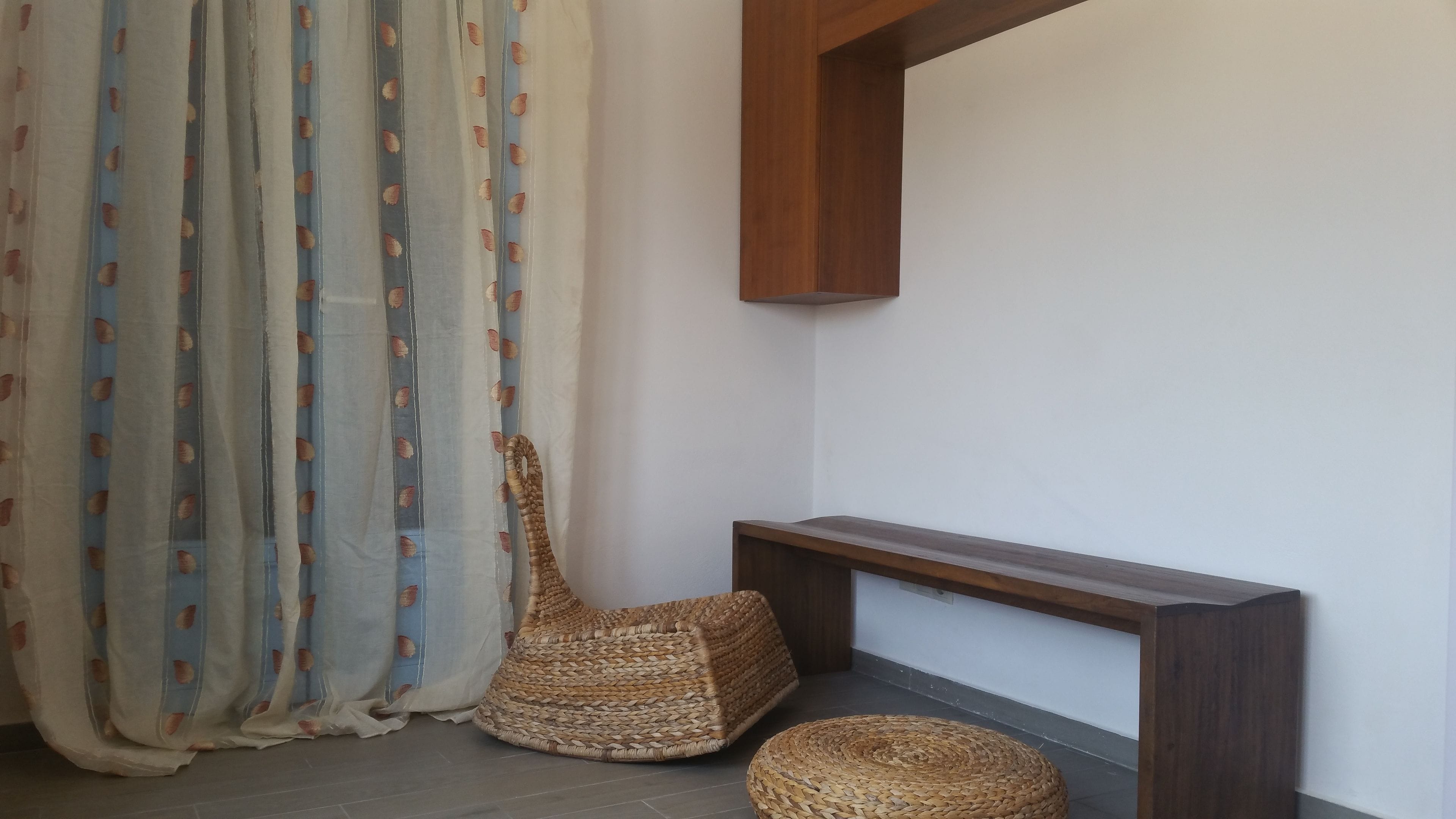 Un coin aménagé avec un fauteuil et un pouf en osier, une console en bois et un rideau léger orné de motifs de feuilles.