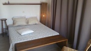 Een slaapkamer met een houten bedframe, een grijs bed met een patroon, twee kussens, een klein krukje en een rieten stoel.