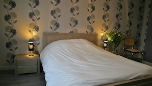 A neatly made bed in a hotel room with floral wallpaper, two bedside lamps, and a small table with a vase of flowers. - Maison d'hotes La Colombelle (Colombé-le-Sec)