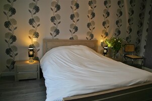 Una cama bien hecha en una habitación de hotel con papel tapiz floral, dos lámparas de noche y una mesita con un jarrón de flores.